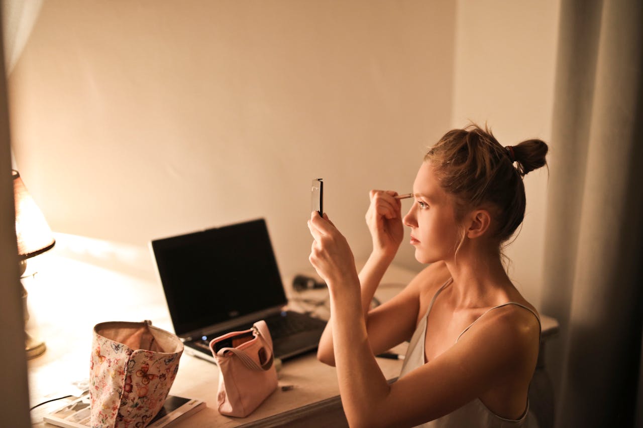 menu-07 A young woman applies makeup at her vanity table, illuminated by soft light, in a cozy home setting.